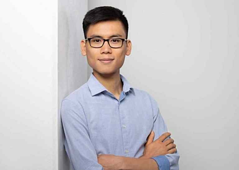 Bewerbungsfotos Berlin - Modernes Bewerbungsfoto eines asiatischen jungen Mannes, mit Brille und blauem Hemd. der locker an einer Säule lehnt vor einem hellen Hintergrund vom Fotostudio Farbtonwerk in Berlin Mitte