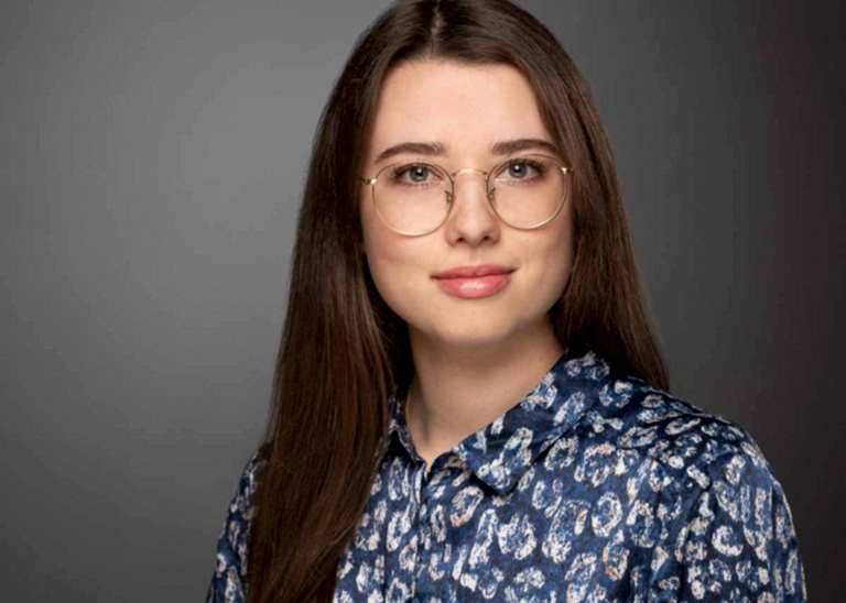 Modernes Bewerbungsfoto einer jungen Frau mit dunkelbraunen, langen Haaren und einer großen Brille, die eine Bluse mit blauem Muster trägt