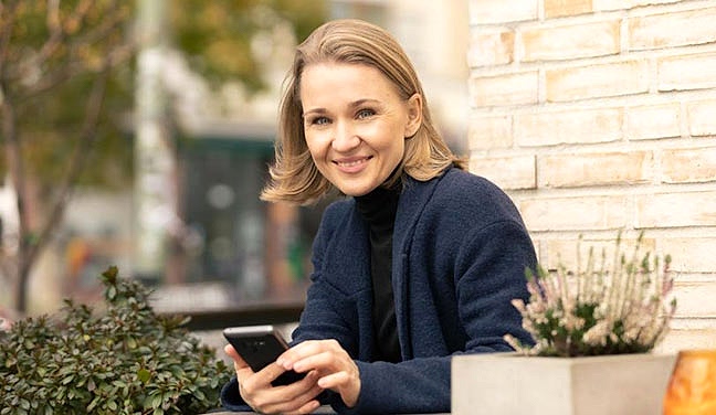 Dating-Foto einer blonden jungen Frau mit schulterlangen Haaren, die mit ihrem Handy in der Hand am Tisch eines Straßenkaffees sitz