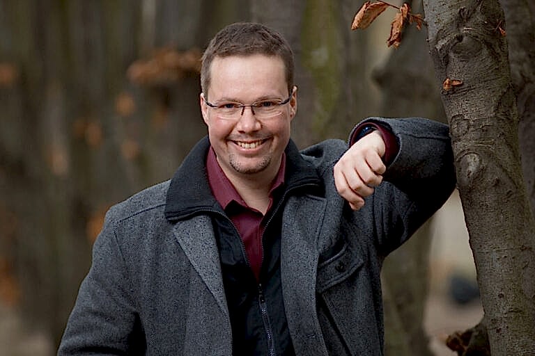 Schauspielerportraits eines dunkelhaarigen Schauspielers mit randloser Brille, der sich in einer grauen Jacke an einen Baum abstützt