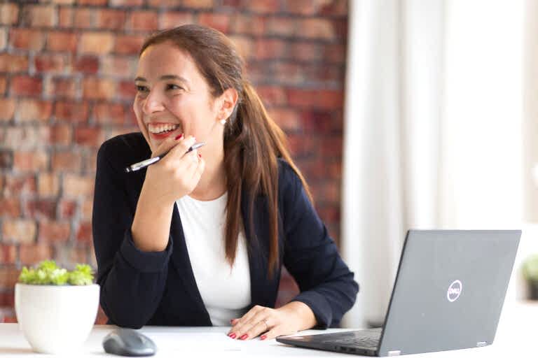 Dating-Foto einer junge, lachenden Frau mit lange, braunen Haaren zu einem Pferdeschwanz zusammengebunden. Sie sitzt mit einem Stift in der Hand in ihrem Büro an einem Schreibtisch mit Laptop und lacht freudig. Im Hintergrund ist eine rohe Backsteinwand.