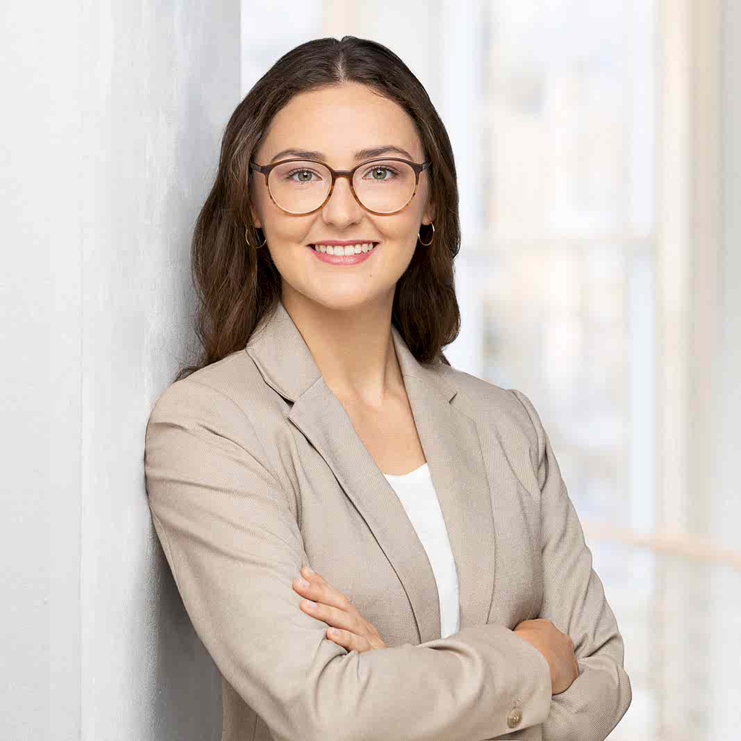 Modernes Profilfoto für Xing und LinkedIn einer Frau mit langen, lockigen Haare und einem beigen Blazer vor einer hellen gewölbten Fesnterfront die entspannt an einer Säule lehnt.